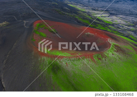 Aerial view of a volcanic crater in Iceland with red volcanic soil, green moss covered slopes, and a barren surrounding landscape with a winding road. Aerial view of a volcanic crater in Iceland with red volcanic soil, green moss covered slopes, and a barren surrounding landscape with a winding road. 133108468