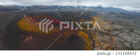 Aerial view of a red and yellow crater in Iceland's volcanic terrain, with snow capped mountains, a winding road, and low hanging clouds in the background. Aerial view of a red and yellow crater in Iceland's volcanic terrain, with snow capped mountains, a winding road, and low hanging clouds in the background. 133108472