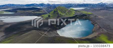 Aerial view of a vivid blue crater lake surrounded by dark volcanic terrain, a green moss covered mountain, and snow capped peaks in Iceland. 133108473