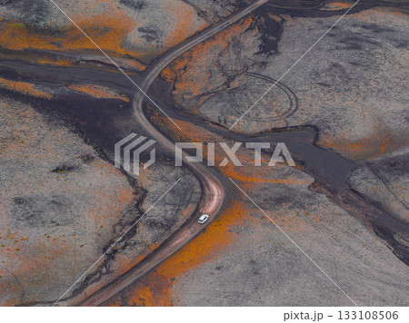 An aerial view shows a dirt road winding through Iceland's volcanic terrain, with dark ashen patches, orange and yellow hues, and a single white vehicle. 133108506