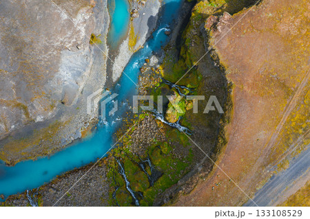 Turquoise river winds through moss covered rocks and earthy terrain in Iceland. Small waterfalls cascade into the river, creating dynamic movement. 133108529