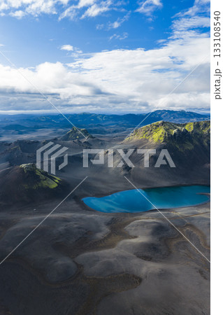 A vivid blue crater lake sits amid dark volcanic terrain in Iceland, with moss covered hills and mountains under soft sunlight and a partly cloudy sky. A vivid blue crater lake sits amid dark volcanic terrain in Iceland, with moss covered hills and mountains under soft sunlight and a partly cloudy sky. 133108540