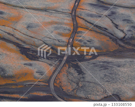 An aerial view of a winding road traversing a barren volcanic terrain in Iceland, featuring dark gray and orange hues from ash and moss or lichen. 133108550