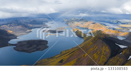 A vast lake in Iceland surrounded by rugged volcanic terrain with yellow and orange hills, deep blue water, and a faint rainbow under a cloudy sky. 133108583
