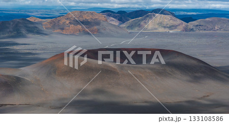 A volcanic crater with reddish and gray tones surrounded by barren ash covered land, multicolored mountains, and a winding road under a cloudy sky. 133108586