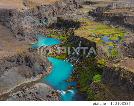 Turquoise waters flow through Sigoldugljufur Canyon in Iceland, with steep rock walls, cascading waterfalls, and green moss contrasting the rugged terrain. 133108587