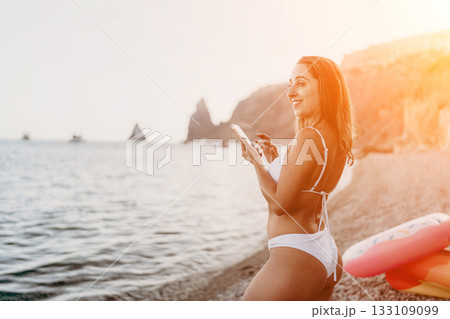 Woman, smartphone, beach. Smiling woman in bikini texts on phone at pebble beach with copy space. 133109099