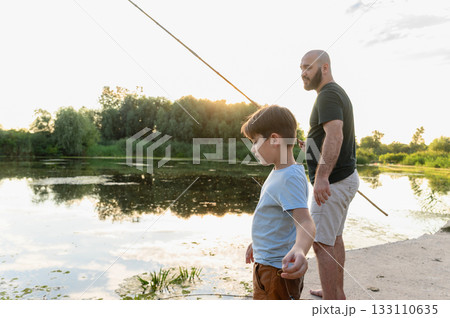 Father and son standing with fishing rods at sunset by river. Father and son standing with fishing rods at sunset by river. 133110635