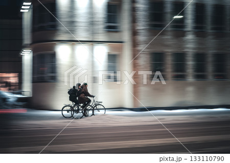 Two people cycling together on snowy city street at night, motion blur capturing winter urban lifestyle and friendship in cold weather Two people cycling together on snowy city street at night, motion blur capturing winter urban lifestyle and friendship in cold weather 133110790