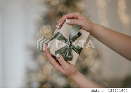 Hands Holding Gift Box with Satin Bow by Christmas Tree 133111462