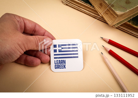 Hand holding card with Greek flag and text learn Greek on beige background Hand holding card with Greek flag and text learn Greek on beige background 133111952