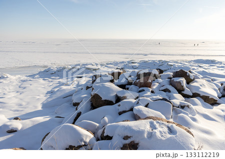 Snow covered rocks dominate the foreground of a quiet, frozen shoreline 133112219