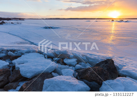 A tranquil winter scene of a frozen sea at sunset, reflecting warm orange light 133112227