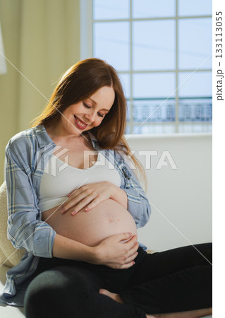 Pregnancy motherhood people expectation future. Pregnant woman touching big belly sitting on couch at home. Girl hugging her tummy enjoying pregnancy. Maternity tenderness parenthood new life concept Pregnancy motherhood people expectation future. Pregnant woman touching big belly sitting on couch at home. Girl hugging her tummy enjoying pregnancy. Maternity tenderness parenthood new life concept 133113055