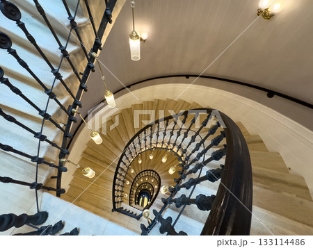 Spiral staircase viewed from above with ornate iron railing and warm hanging lights in historic interior 133114486