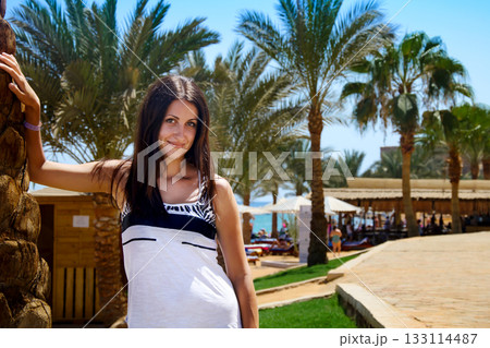 oung girl with long dark hair with in the white T-shirt posing against the backdrop of palm trees on a sunny day. 133114487