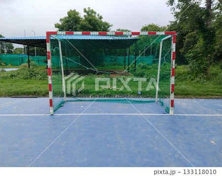 Outdoor futsal goalpost with striped metal frame and loose green net on blue sports court, standalone mini football goal in open ground surrounded by grass and trees, dedicated small-sided play area Outdoor futsal goalpost with striped metal frame and loose green net on blue sports court, standalone mini football goal in open ground surrounded by grass and trees, dedicated small-sided play area 133118010
