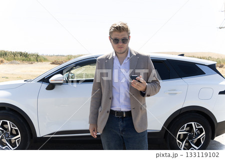 A man is standing next to a white car while holding a small device A man is standing next to a white car while holding a small device 133119102