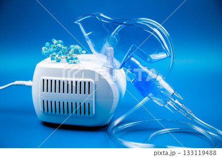 The blue breathing mask of the inhaler, from which steam is emitted, lies on the device on a blue background. The concept of treatment of respiratory tract diseases, lung diseases, medicines. High The blue breathing mask of the inhaler, from which steam is emitted, lies on the device on a blue background. The concept of treatment of respiratory tract diseases, lung diseases, medicines. High 133119488