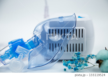 Close-up of the breathing blue mask of the inhaler lying against the background of the device, next to it is an ampoule with medicine and flowers on a white background. The concept of treating 133119524