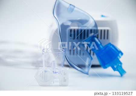 A close-up of an inhalation bowl and an ampoule with a medicinal solution stands against the background of a blue breathing mask and a device on a white background. The concept of treatment of 133119527