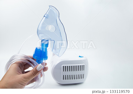 Close-up side view of a blue inhalation mask held in the hand against the background of a white device on a white background. The concept of inhalation of vapors, treatment of the respiratory tract Close-up side view of a blue inhalation mask held in the hand against the background of a white device on a white background. The concept of inhalation of vapors, treatment of the respiratory tract 133119570