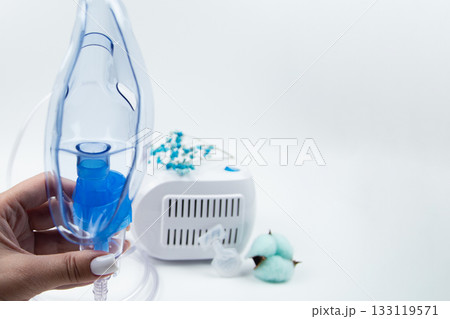 Close-up from the side of a blue mask for inhalation, holding a white device in out-of-focus with blue flowers on a white background. The concept of inhalation of vapors, treatment of the respiratory Close-up from the side of a blue mask for inhalation, holding a white device in out-of-focus with blue flowers on a white background. The concept of inhalation of vapors, treatment of the respiratory 133119571