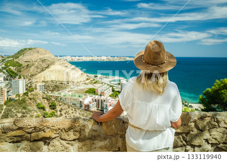 Woman in a hat looking at the blue sea and city from the viewpoint in Alicante, Spain, on a sunny summer day. 133119940
