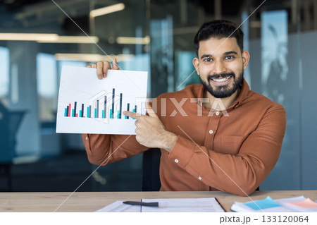 Smiling young indian businessman holding up a graph and pointing, explaining financial statistics during a presentation in a modern office setup, analyzing market trends Smiling young indian businessman holding up a graph and pointing, explaining financial statistics during a presentation in a modern office setup, analyzing market trends 133120064