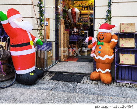 Inflatable Santa Claus and gingerbread man decorations stand outside a shop entrance. Santa, in his traditional red outfit, waves with one hand Inflatable Santa Claus and gingerbread man decorations stand outside a shop entrance. Santa, in his traditional red outfit, waves with one hand 133120484