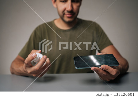 Blurred portrait of man spraying sanitizer onto mobile phone screen, highlighting importance of hygiene and disinfection in everyday life on gray table surface. Concept of phone maintenance Blurred portrait of man spraying sanitizer onto mobile phone screen, highlighting importance of hygiene and disinfection in everyday life on gray table surface. Concept of phone maintenance 133120601