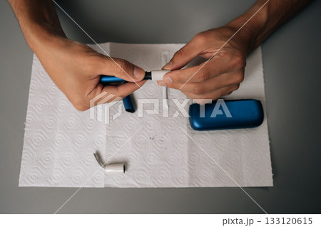 Top view of unrecognizable man cleaning electronic cigarette with cleaning stick and paper on table, performing maintenance, ensuring proper hygiene, sanitizing personal vaping equipment. Top view of unrecognizable man cleaning electronic cigarette with cleaning stick and paper on table, performing maintenance, ensuring proper hygiene, sanitizing personal vaping equipment. 133120615