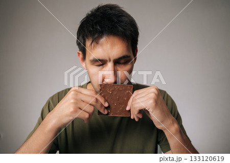 Studio portrait of enjoying young man savoring rich aroma of dark chocolate bar with closed eyes, holding it close to face in anticipation of delightful treat, sitting at table on isolated background. Studio portrait of enjoying young man savoring rich aroma of dark chocolate bar with closed eyes, holding it close to face in anticipation of delightful treat, sitting at table on isolated background. 133120619