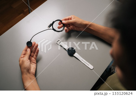 Top view from shoulder of unrecognizable man holding smartwatch inductive charger, preparing to power up smart watch, sitting at table. Concept of healthy lifestyle. 133120624