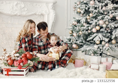 Blonde mother, brunette father and little daughter sitting on a floor near Christmas tree. Woman and daughter wearing plaid pajamas. Family opening a Christmas gifts together. Blonde mother, brunette father and little daughter sitting on a floor near Christmas tree. Woman and daughter wearing plaid pajamas. Family opening a Christmas gifts together. 133122556