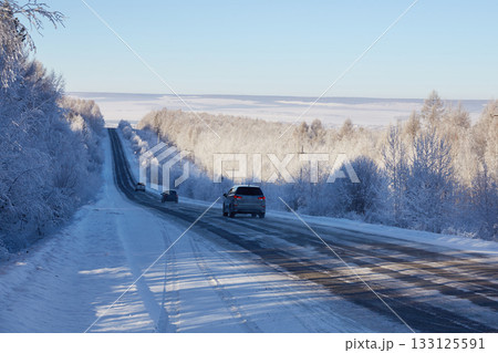 Cars drive along an asphalt road among a snowy winter forest 133125591