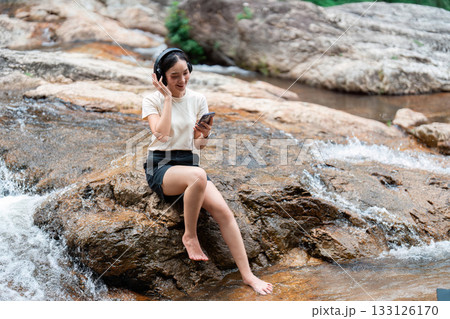 Relaxation and Wellness. Young woman enjoying music by a serene river. 133126170
