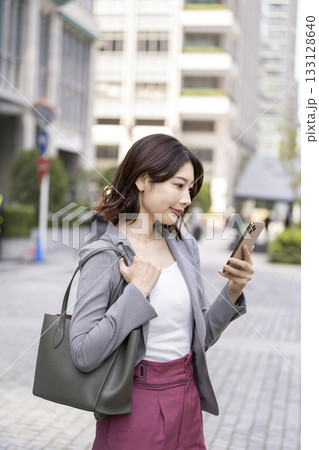 オフィス街でスマートフォンの画面を見るビジネスウーマン 133128640