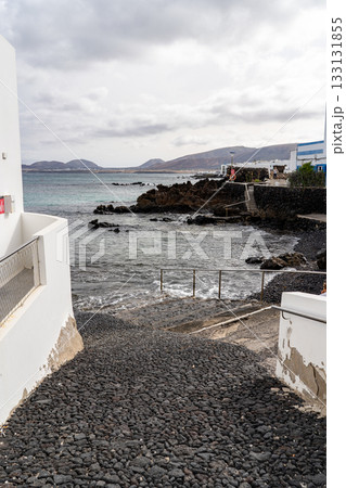 A view of a rocky coast in Punta Mujeres, Lanzarote. The water gently laps against the shore with buildings in view. 133131855