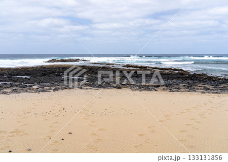 A serene beach scene on La Graciosa, with waves crashing against dark volcanic rocks and sandy shore. Footprints mark the sand. 133131856