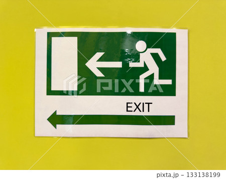 Green sign on yellow wall with building exit direction. Safe navigation indoors in case of fire or accident. Street exit sign with directional arrow and symbol of person running out Green sign on yellow wall with building exit direction. Safe navigation indoors in case of fire or accident. Street exit sign with directional arrow and symbol of person running out 133138199