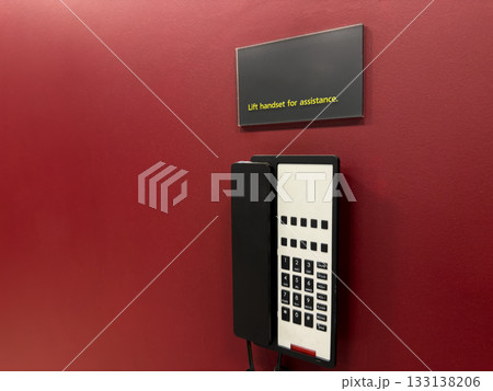 Landline telephone on red wall with warning sign directing you to operator. Emergency communication within building in case of problems or accidents. Phone with communication buttons Landline telephone on red wall with warning sign directing you to operator. Emergency communication within building in case of problems or accidents. Phone with communication buttons 133138206