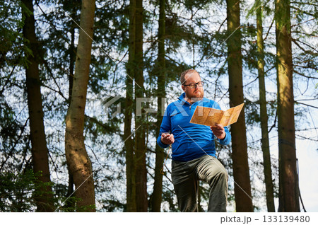 Man traveler in glasses stands in dense forest, holding map in one hand and compass in other. Tourist planning outdoor adventure or hiking route, amidst tall trees and natural surroundings. Man traveler in glasses stands in dense forest, holding map in one hand and compass in other. Tourist planning outdoor adventure or hiking route, amidst tall trees and natural surroundings. 133139480