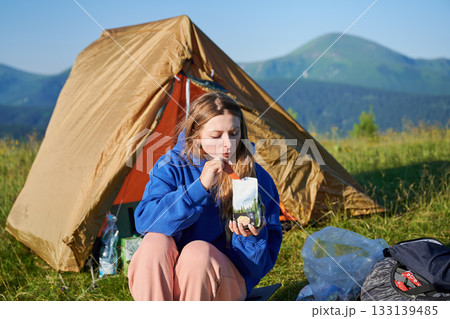 Young woman enjoying meal from food pouch. Tourist in blue hoodie and pink pants sits near orange tent, uses spoon with serene mountainous background. Young woman enjoying meal from food pouch. Tourist in blue hoodie and pink pants sits near orange tent, uses spoon with serene mountainous background. 133139485