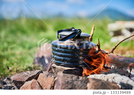 Close-up of blackened kettle heating over campfire, with flames dancing around it. Kettle on ring of stones in grassy meadow. On blurred background natural, outdoor setting with hint of mountains. 133139489