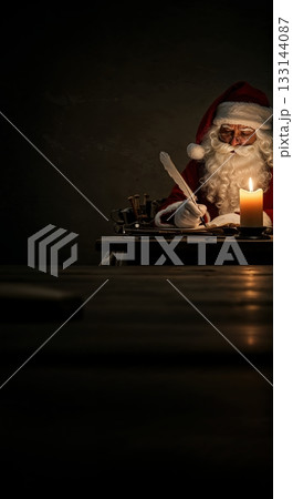 An elderly man resembling Santa Claus in a red suit and white beard meticulously writes with a quill pen by warm candlelight on a rustic wooden table. 133144087