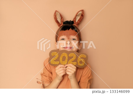 A funny girl dressed up in a fire horse costume holds the golden numbers 2026 in hands. The symbol of the new year according to the Eastern calendar 133146099