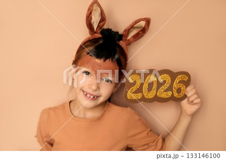 A cute sweet child in a fire horse costume with a painted face, with ear-shaped headbands, shows big numbers 2026. The symbol of the new year 133146100