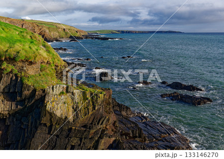 View of the ocean waves crashing against the rocky cliffs along the coastline. Lush green grass covers the top of the cliffs in West Cork, Ireland, on a partly cloudy day. 133146270