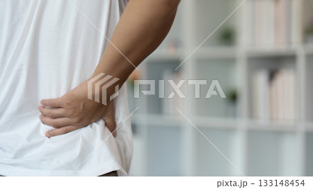 Back Pain Awareness. Young man holding lower back in discomfort while seated. Back Pain Awareness. Young man holding lower back in discomfort while seated. 133148454